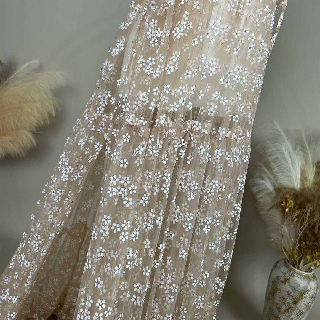 A khaki tulle dress with white floral embroidery and gold glitter accents, displayed against a neutral backdrop with dried floral arrangements.