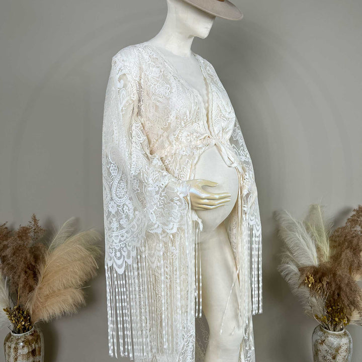 A beige lace maternity with long sleeves and fringe trim, displayed on a pregnant mannequin. 
