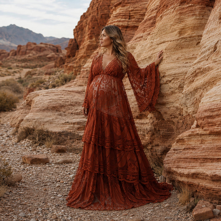 A pregnant woman stands on a gravel path next to layered red rock formations in a canyon landscape, wearing a terracotta tiered lace maternity dress. 