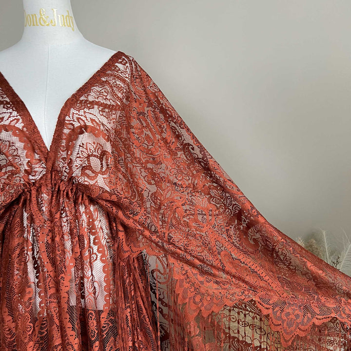 A mannequin displays a close-up of a terracotta lace cape featuring floral openwork texture and a deep V-neck; the fabric is semi-transparent mesh.