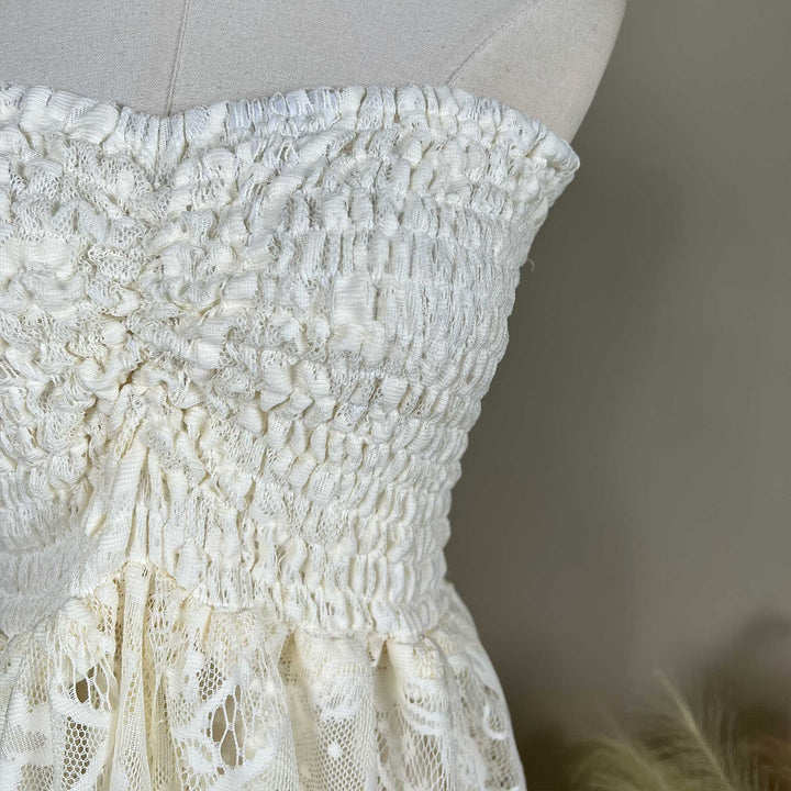 A mannequin displays a close-up of a beige strapless dress bodice with dense shirring and lace fabric; the waist connects to a tiered skirt.