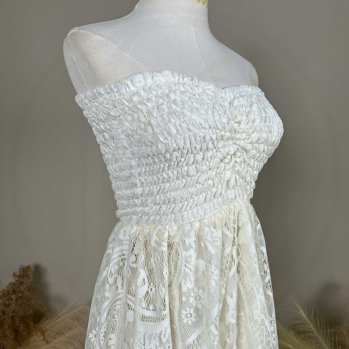 A mannequin displays a close-up of a beige strapless dress bodice with dense shirring and lace fabric; the waist connects to a tiered skirt, set against a solid wall.