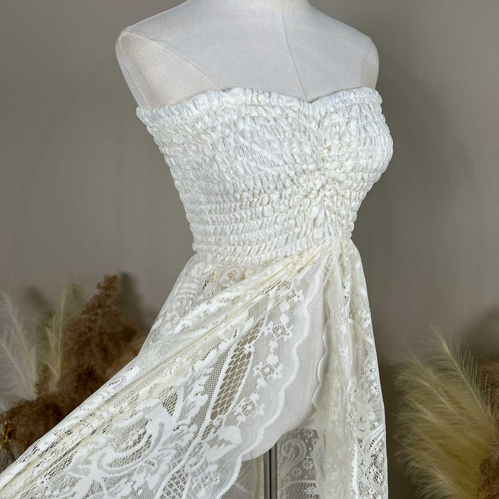 A mannequin displays a close-up of a beige strapless dress bodice with dense shirring and lace fabric; the waist connects to a tiered skirt, set against a solid wall.