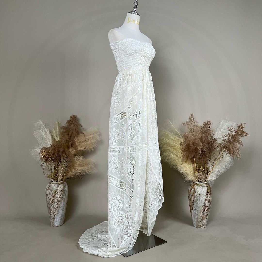 A mannequin shows a side view of a beige strapless lace gown with a ruched bodice and train; the dress features an openwork pattern, set against a gray wall with dried flower vases on both sides.