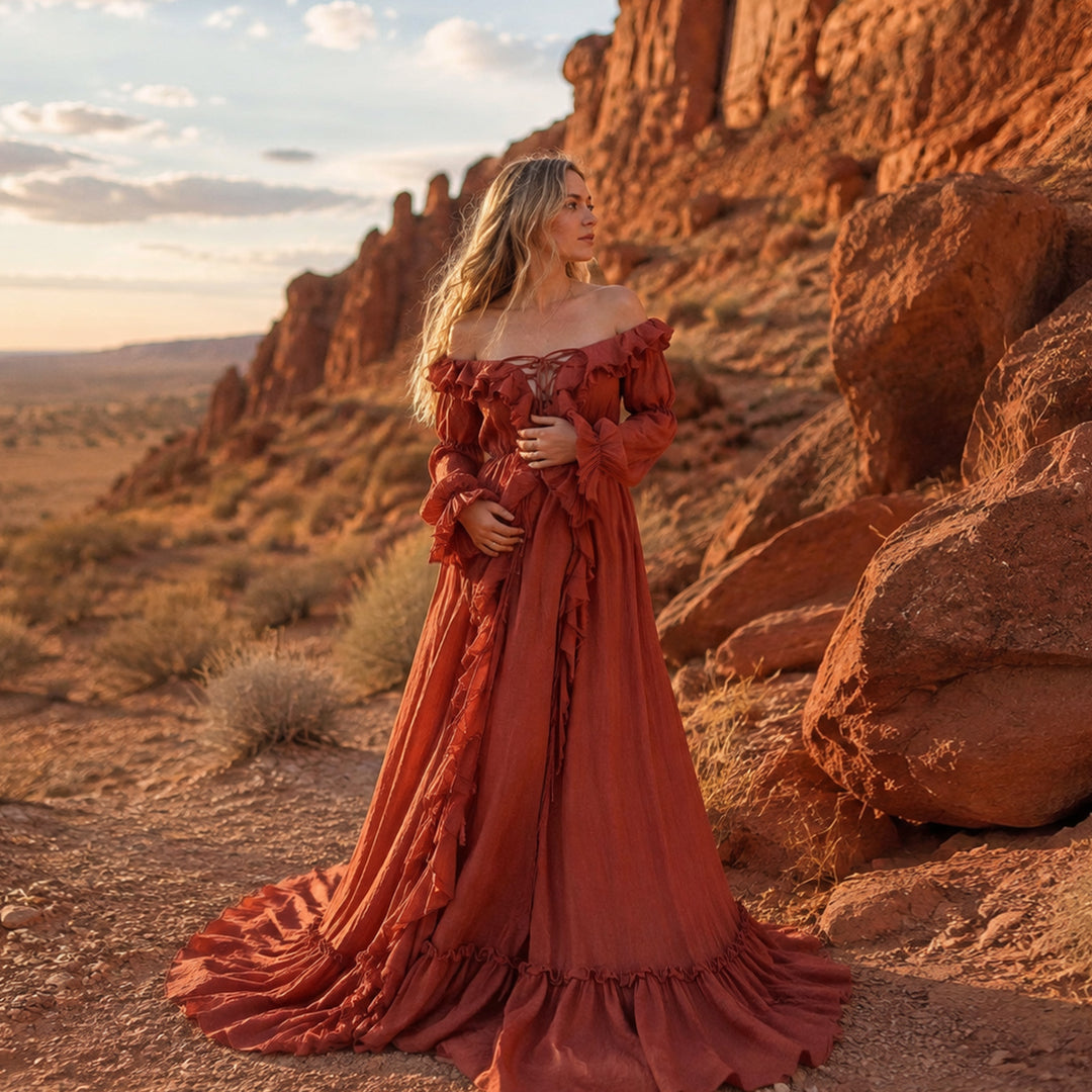 A woman stands outdoors in a desert canyon wearing a long, off-the-shoulder kallectoria terracotta 
gown with ruffled details and a full skirt 