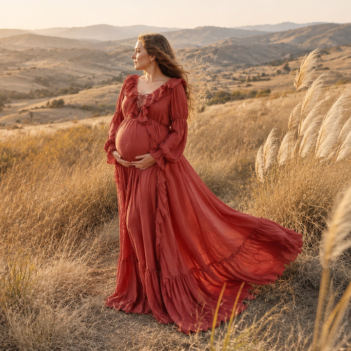 A pregnant woman stands in a dry grassy landscape wearing a flowing terracotta kallectoria maternity gown with long ruffled sleeves and a full skirt that spreads across the ground. 