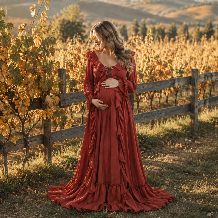 A pregnant woman stands in a vineyard wearing a long-sleeved terracotta kallectoria dress with ruffle details.