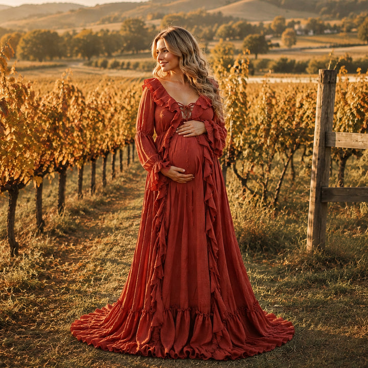 A pregnant woman wearing a long-sleeved terracotta ruffled kallectoria gown stands in a vineyard at sunset, holding her baby bump. 