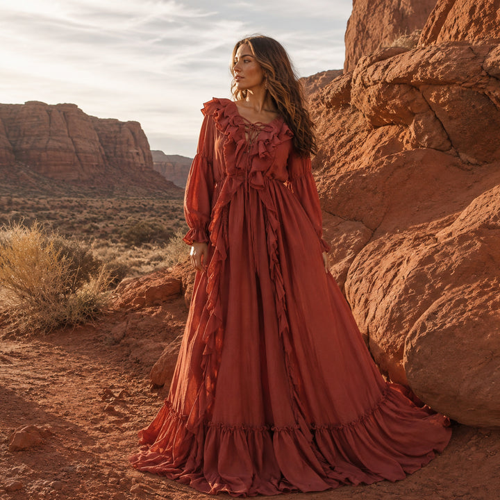 A woman wearing a long, ruffled kallectoria dress in a terracotta color stands outdoors in a desert canyon setting with large red rock formations and dry grasses in the background.