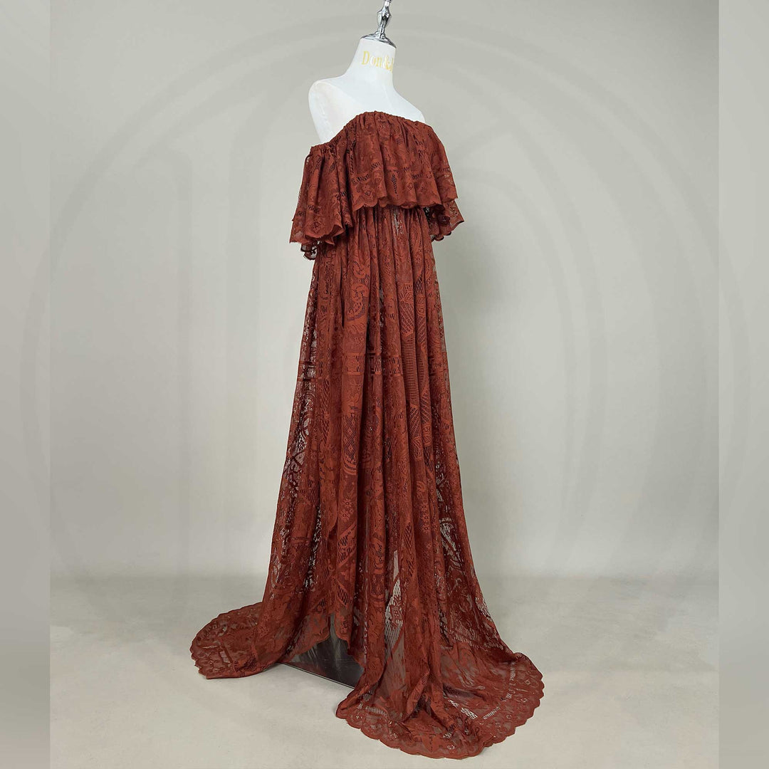 A mannequin a side view of a terracotta off-shoulder lace gown with ruffled sleeves and a train; the dress features a sheer openwork structure, set against a gray wall with dried flower vases on both sides.