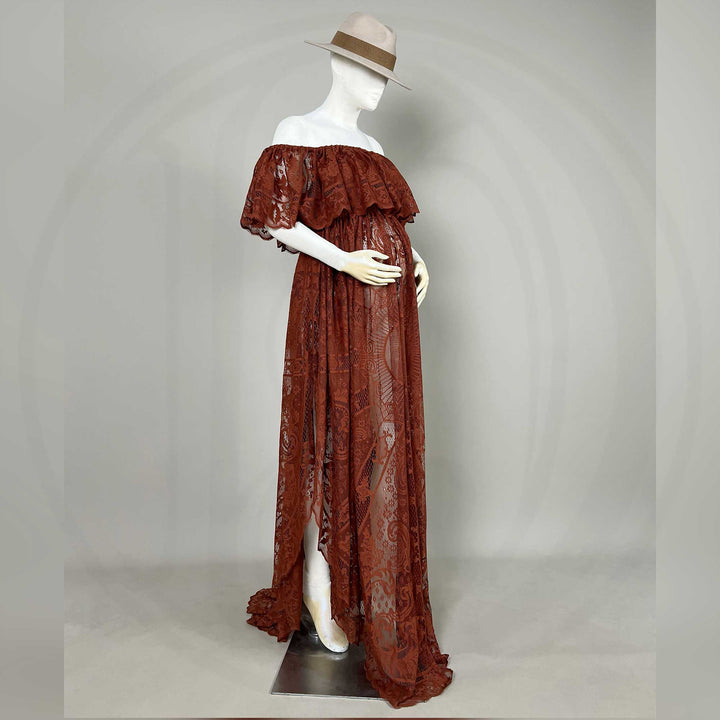 A hat-wearing pregnant mannequin a side view of a terracotta off-shoulder lace gown with ruffled sleeves and a train; the dress features a sheer openwork structure, set against a gray wall with dried flower vases on both sides.