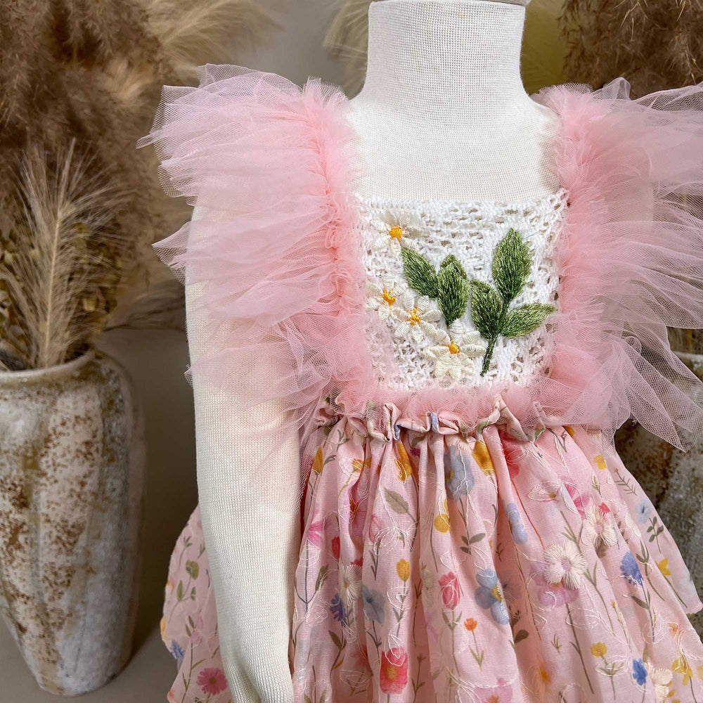A close-up of a pink floral dress with a crochet bodice, green leaf embroidery, and tulle sleeves; the skirt features layered tulle.