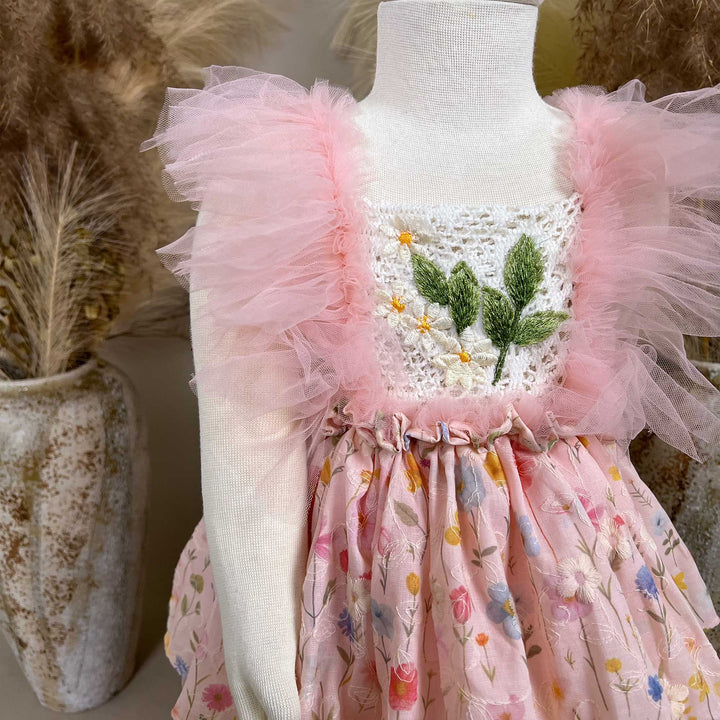A close-up of a pink floral dress with a crochet bodice, green leaf embroidery, and tulle sleeves; the skirt features layered tulle.