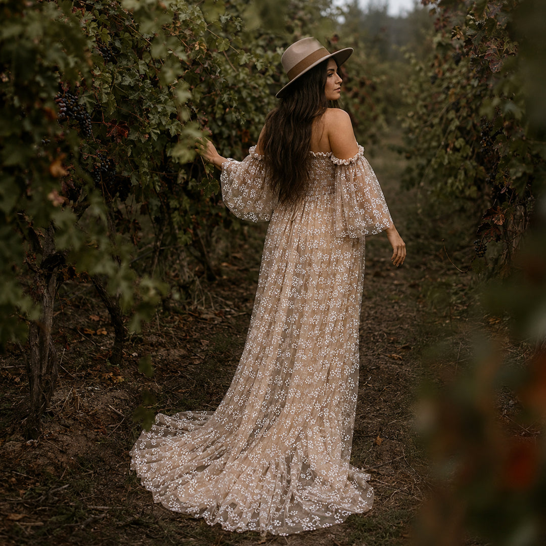 A pregnant woman stands in a vineyard aisle wearing a wide-brimmed hat and a khaki off-shoulder floral gown. 