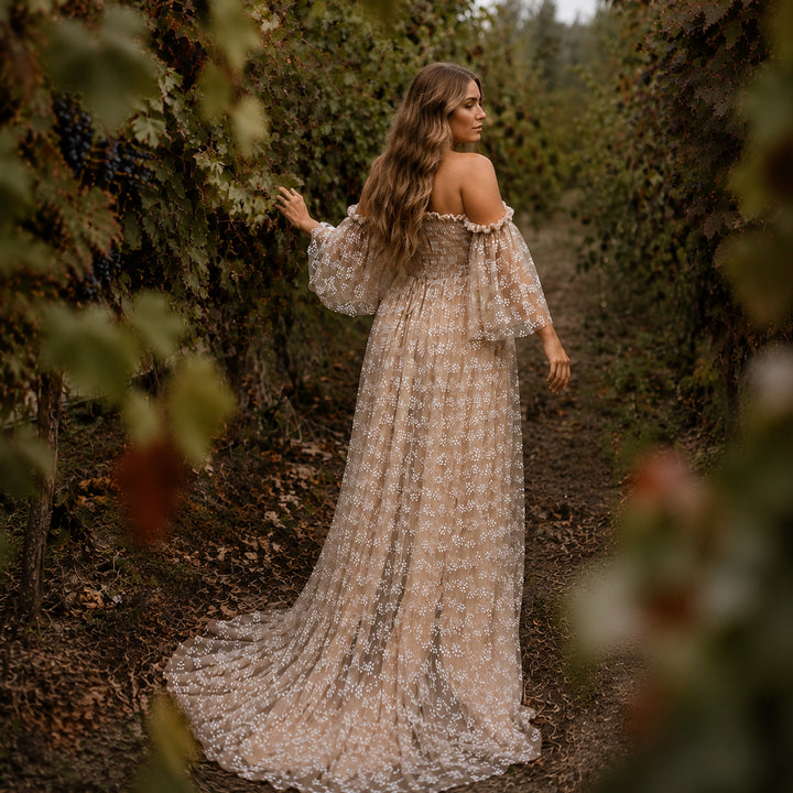 A pregnant woman walks down a path in a vineyard, surrounded by grapevines, wearing a Lysianassa khaiki floor-length khaki tulle dress with floral embroidery. 