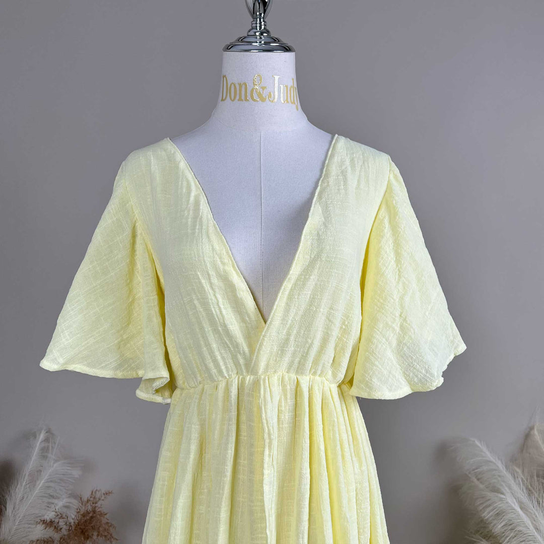 A mannequin displays the front of a yellow V-neck short-sleeve dress, showing a deep-V cut and waist pleats; the fabric has a textured finish.