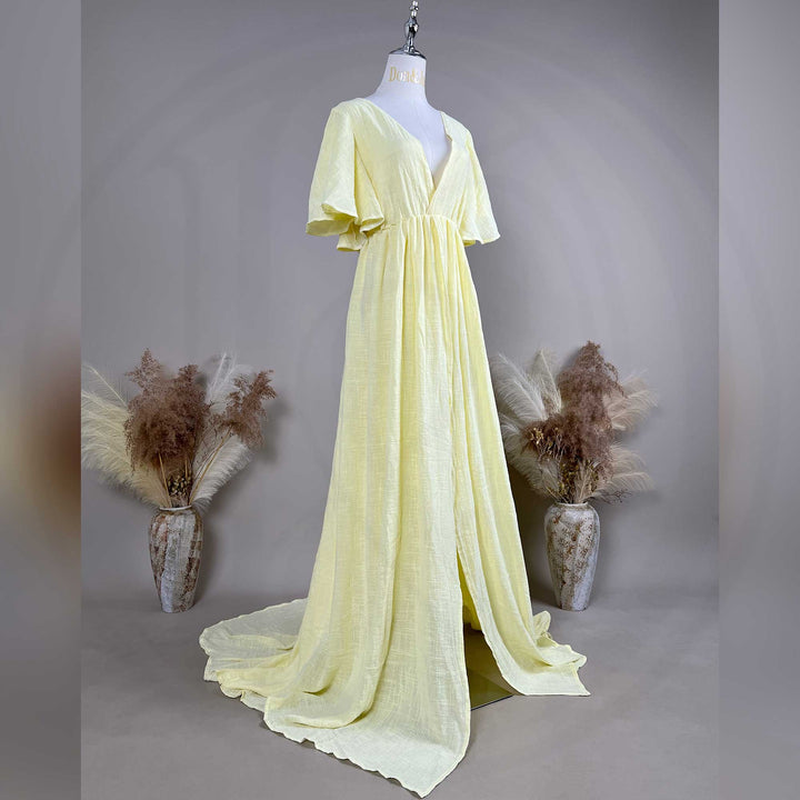 A mannequin shows a side view of a yellow V-neck short-sleeve gown with a high-waist pleat and train; the fabric has a textured finish, set against a solid wall with dried flower vases on both sides.