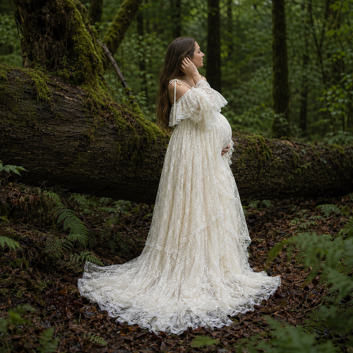A pregnant woman stands in forest landscape wearing a flowing ivory lace maternity gown with off the shoulder ruffled sleeves and a long, tiered skirt that spreads across the leafy ground.