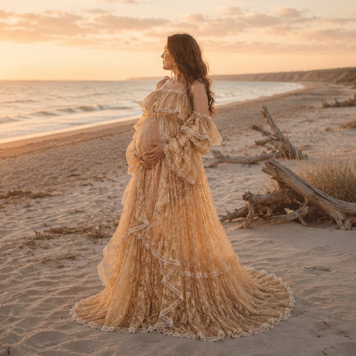 A pregnant woman stands on a sandy beach at sunset, wearing a flowing lace maternity gown with off the shoulder ruffled sleeves and a long, tiered skirt that spreads across the ground