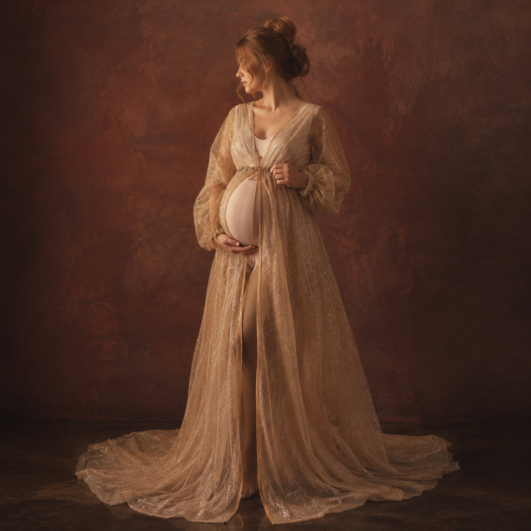 A pregnant model in a khaki sequined V-neck robe stands against a dark red background, wearing a light bodysuit underneath with one hand on her belly; the robe features puff sleeves, a tie belt, and a floor-length train.