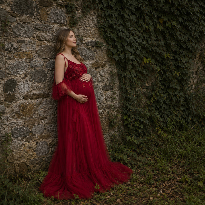 A pregnant woman wears a Oreades red dress with 3D floral embroidery and ruffled sleeves, it's train trailing on the ground; she hold her belly with her right, standing.