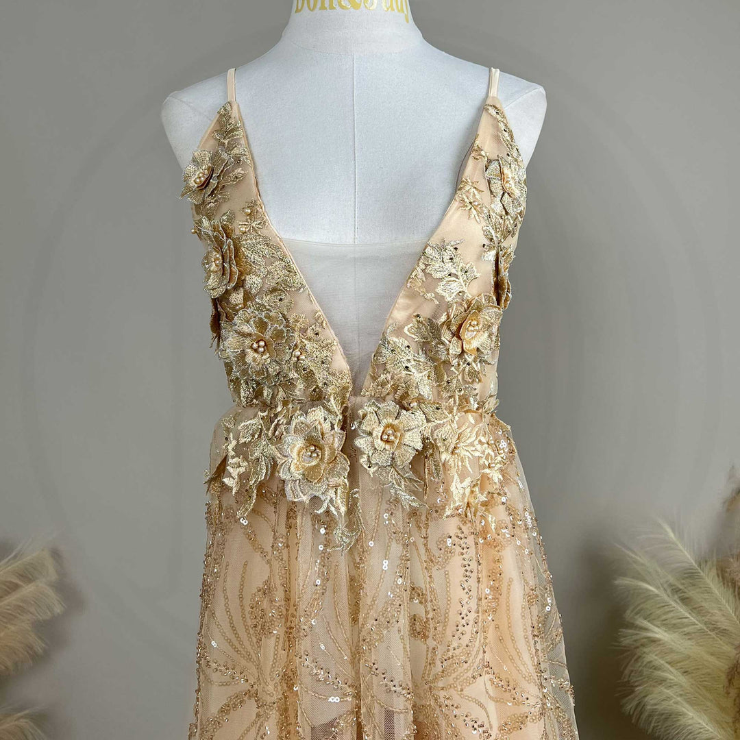 A mannequin displays the upper body of a gold spaghetti strap gown with a deep V-neck and 3D floral decorations.