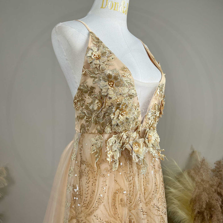 A mannequin displays the upper body of a gold spaghetti strap gown with a deep V-neck and 3D floral decorations.