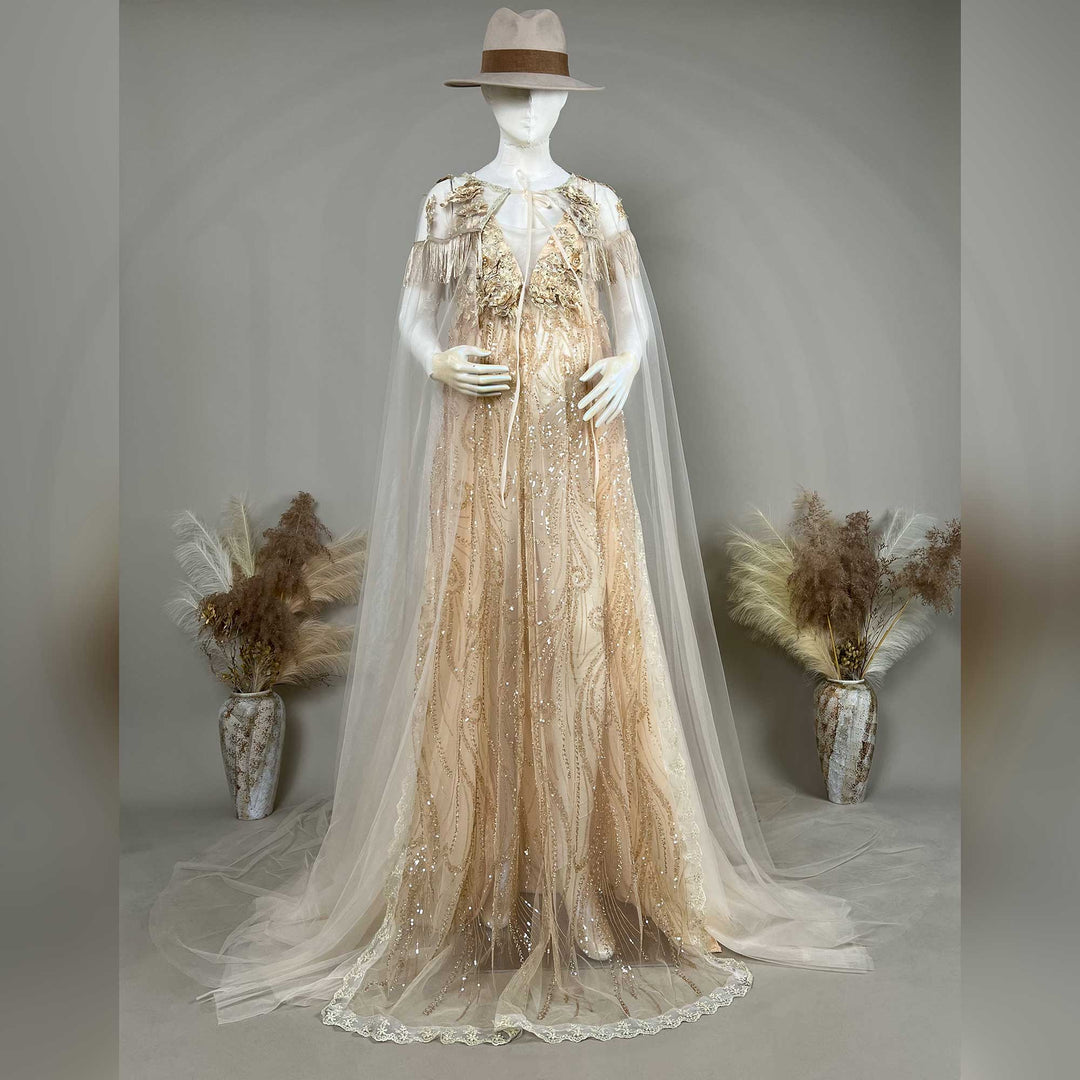 A hat-wearing mannequin displays a gold sequined gown with a tulle cape, featuring fringe cuffs and a train skirt.