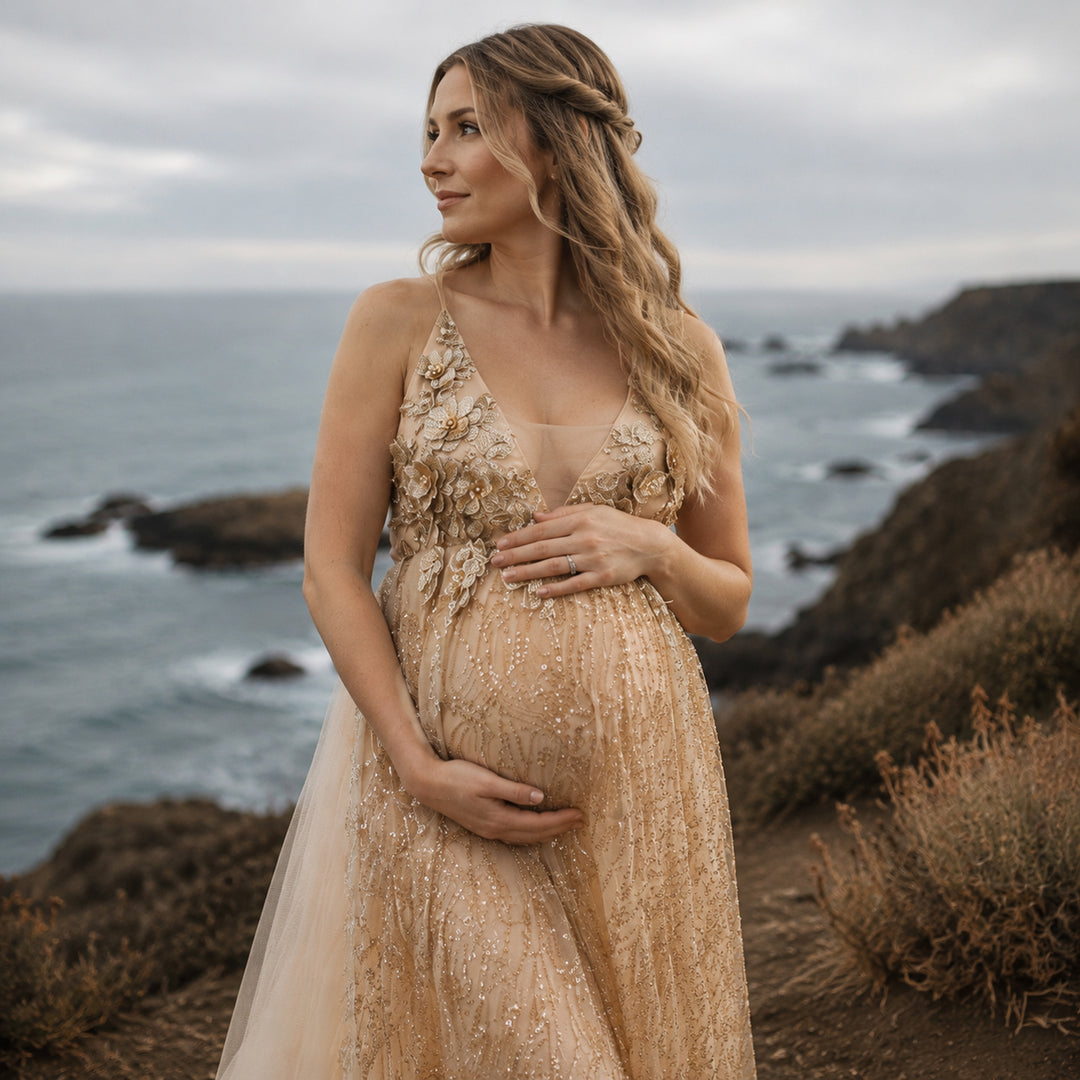 A pregnant model in a gold sequined spaghetti strap gown stands on a seaside cliff, hands on belly.