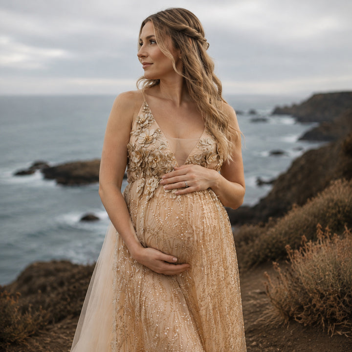 A pregnant model in a gold sequined spaghetti strap gown stands on a seaside cliff, hands on belly.