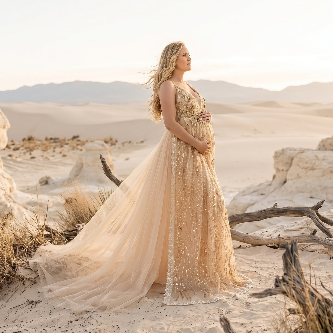 A pregnant model in a gold sequined spaghetti strap gown stands in the desert, hands on belly.