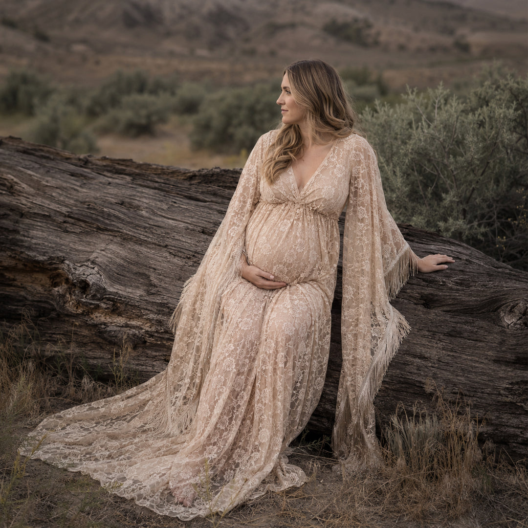 A pregnant woman in a beige lace fringe gown sits sideways on wilderness deadwood, hands on belly.