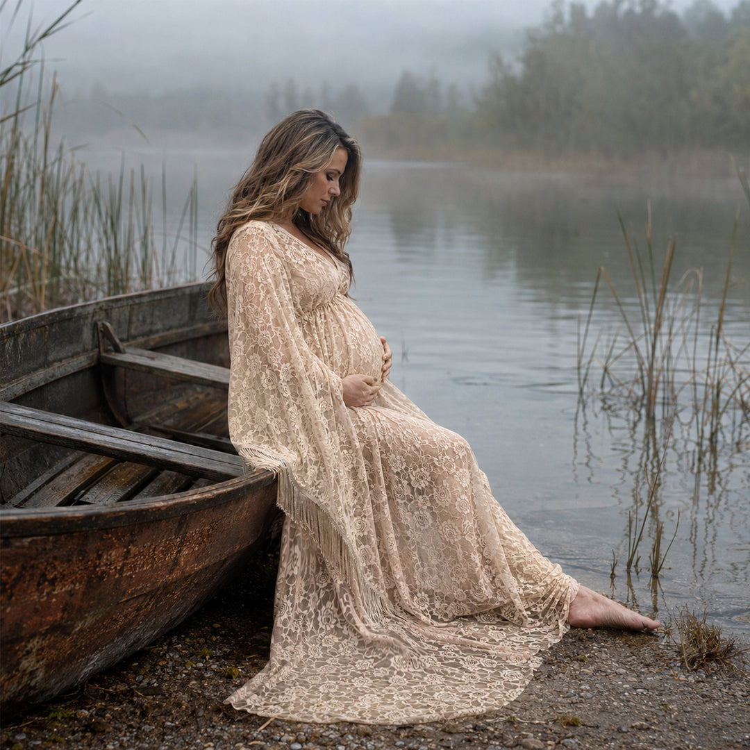 A pregnant woman in a khaki lace fringe gown sits by an old wooden boat at a lakeside, hands on belly.