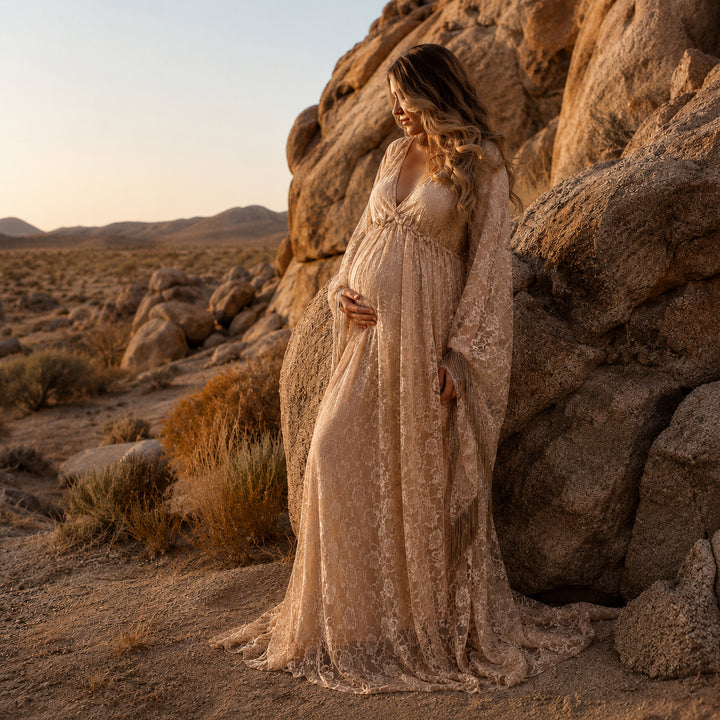 A pregnant woman in a khaki lace fringe gown stands beside desert boulders, one hand on belly.