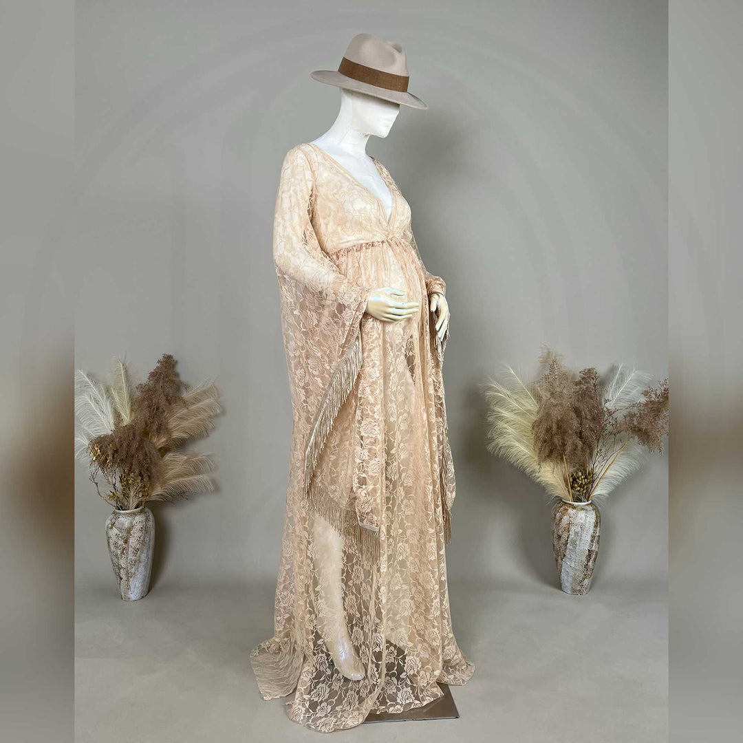 A pregnant mannequin wearing a hat displays a khaki lace gown, featuring a V-neck, fringe sleeves, and a floor-sweeping skirt.