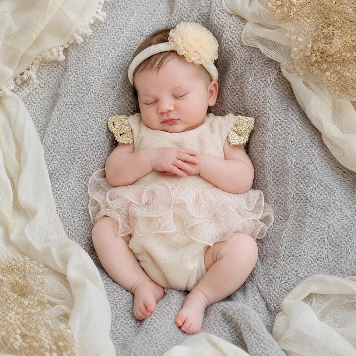 A newborn lies supine on a gray knitted blanket, wearing a skin polar fleece romper with crocheted short sleeves and a multi ayered organza ruffled skirt hem. 