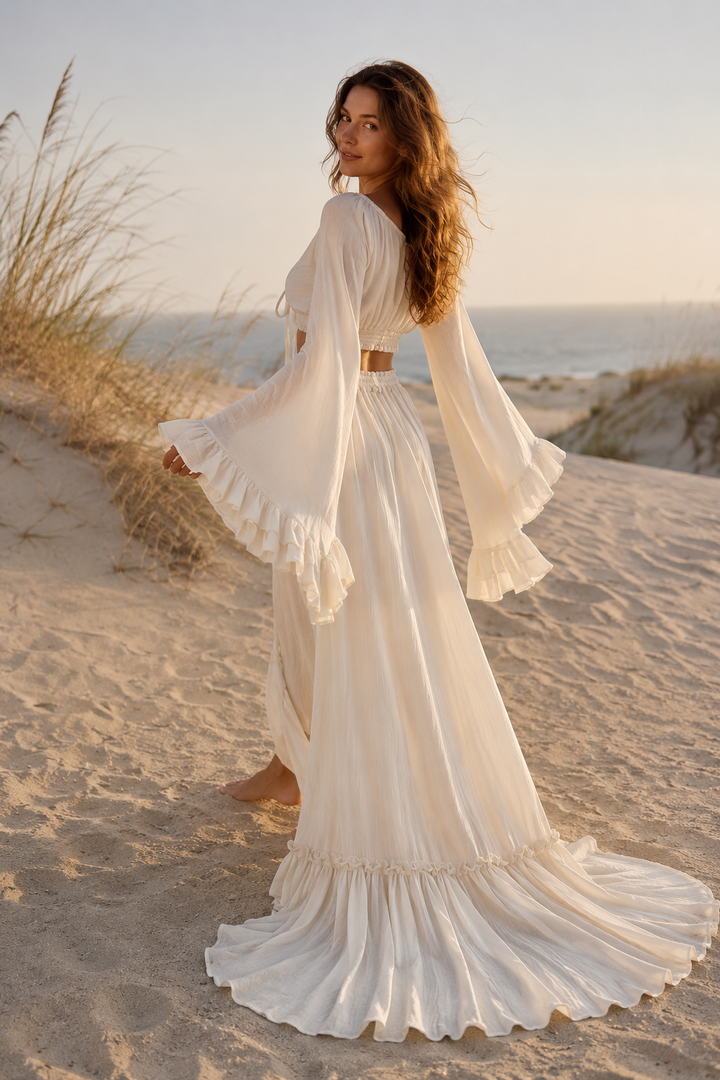 A woman stands sideways on sand in a  natural off white two-piece gown; the top features long boho sleeves with tiered ruffled flares, and the skirt is a high waisted floor length style with a ruffled hem.