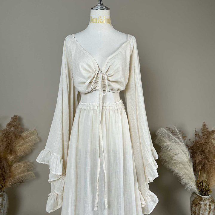 A natural off white two-piece gown is displayed on a mannequin; the top is a deep V boho crop with tiered trumpet sleeves, and the skirt is a high-waisted elastic waist style with a ruffled hem.