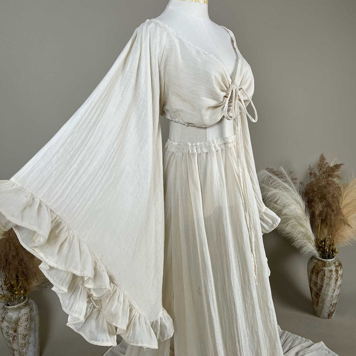 A natural off white two-piece gown is displayed on a mannequin; the top is a deep-V boho crop with tiered trumpet sleeves, and the skirt is a high-waisted elastic-waist style with a ruffled hem.