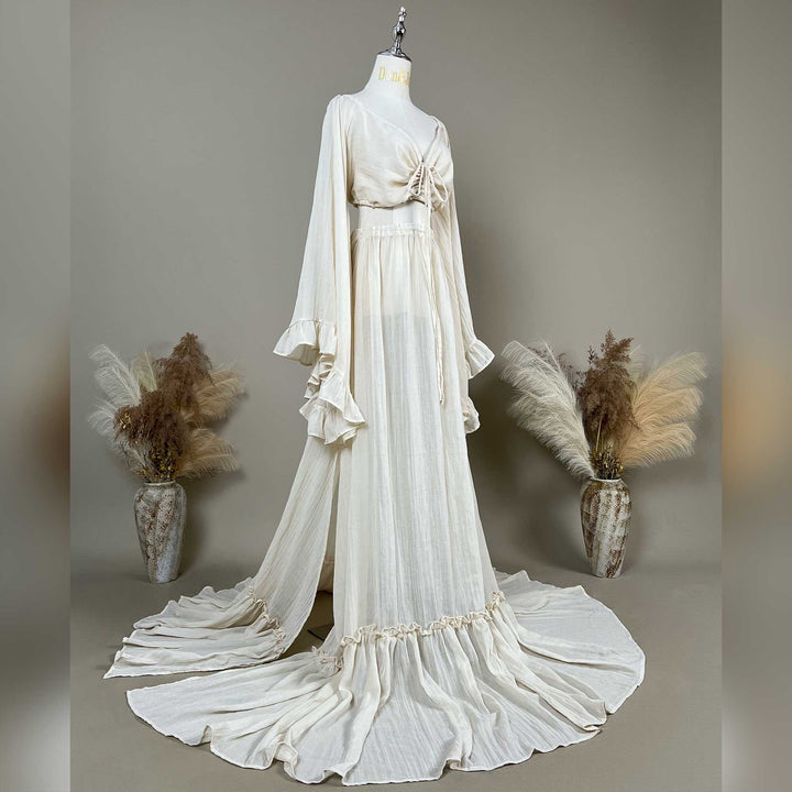 A natural off white two-piece gown is displayed on a mannequin; the top is a deep-V boho crop with tiered trumpet sleeves, and the skirt is a high-waisted floor-length style with a ruffled hem.