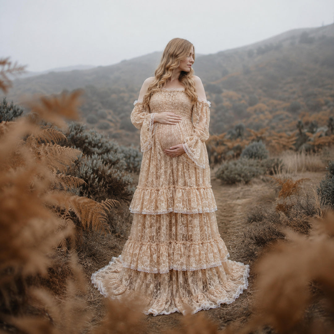 A pregnant woman in a khaki lace off-shoulder gown stands in the wild; the dress features bell sleeves, a tiered ruffled hem, and a train. She cradles her belly with both hands.