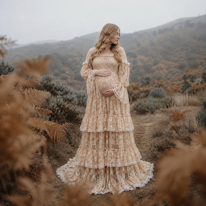 A pregnant woman in a khaki lace off-shoulder gown stands in the wild; the dress features bell sleeves, a tiered ruffled hem, and a train. She cradles her belly with both hands.