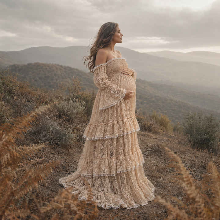 A pregnant woman in a khaki lace off-shoulder gown stands in the wild; the dress features bell sleeves, a tiered ruffled hem, and a train. She cradles her belly sideways with both hands.