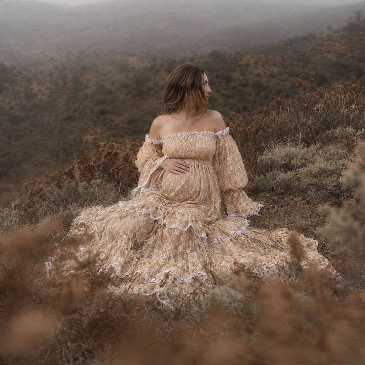 A pregnant woman in a khaki lace off-shoulder gown sits in the wild; the dress features lantern sleeves, a tiered ruffled hem, and a train. She cradles her belly sideways with one hand.