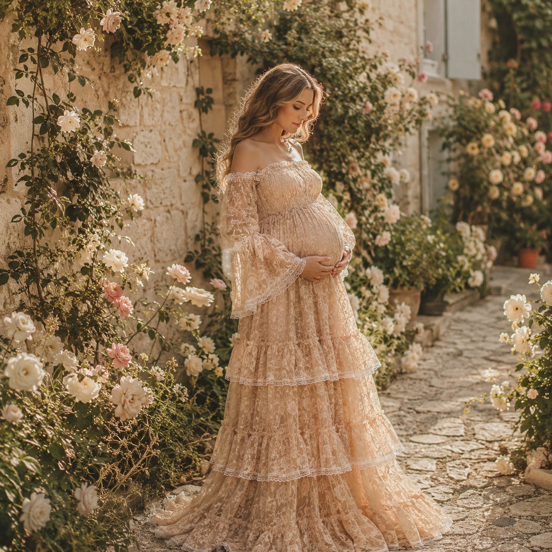A pregnant woman in a khaki Selene off-shoulder lace dress stands on a stone path surrounded by flowers. 