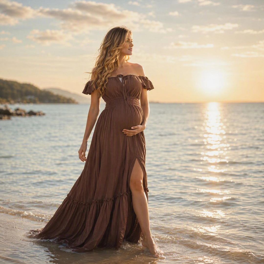 A pregnant woman in a coffee off-shoulder gown stands in shallow sea water; the dress has ruffled sleeves, a front tie, elasticated waist, side slit, and tiered hem. 