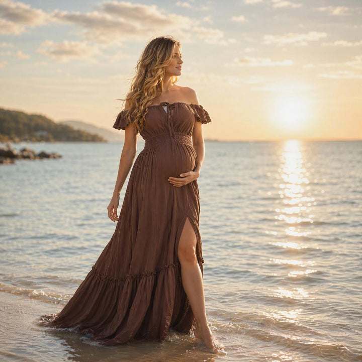 A pregnant woman in a coffee off-shoulder gown stands in shallow sea water; the dress has ruffled sleeves, a front tie, elasticated waist, side slit, and tiered hem. 