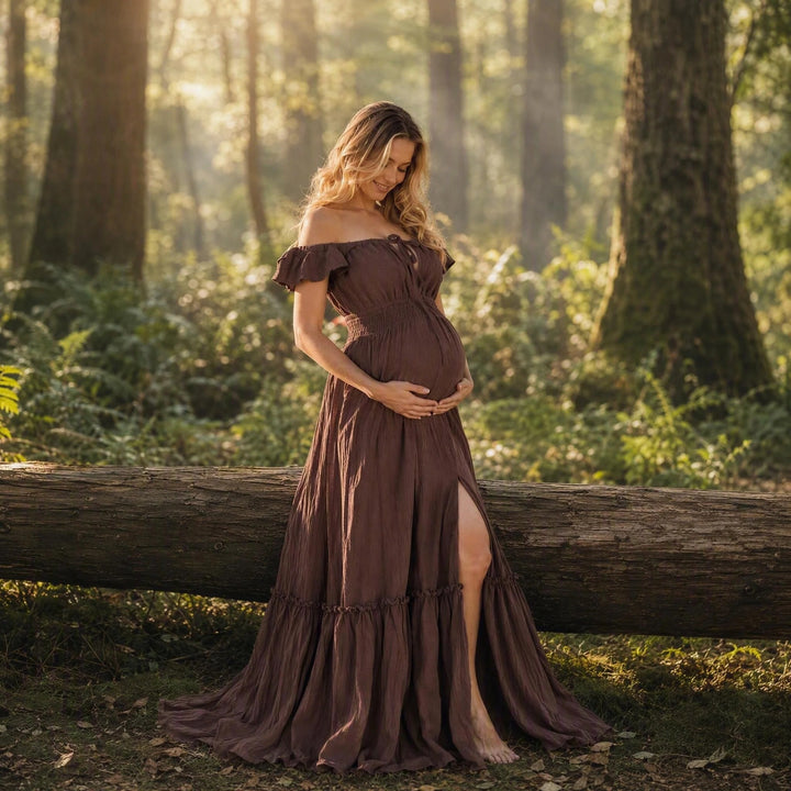 A pregnant woman in a coffee off-shoulder gown stands before a log in a forest; the dress has ruffled sleeves, a front tie, elasticated waist, side slit, and tiered hem. 