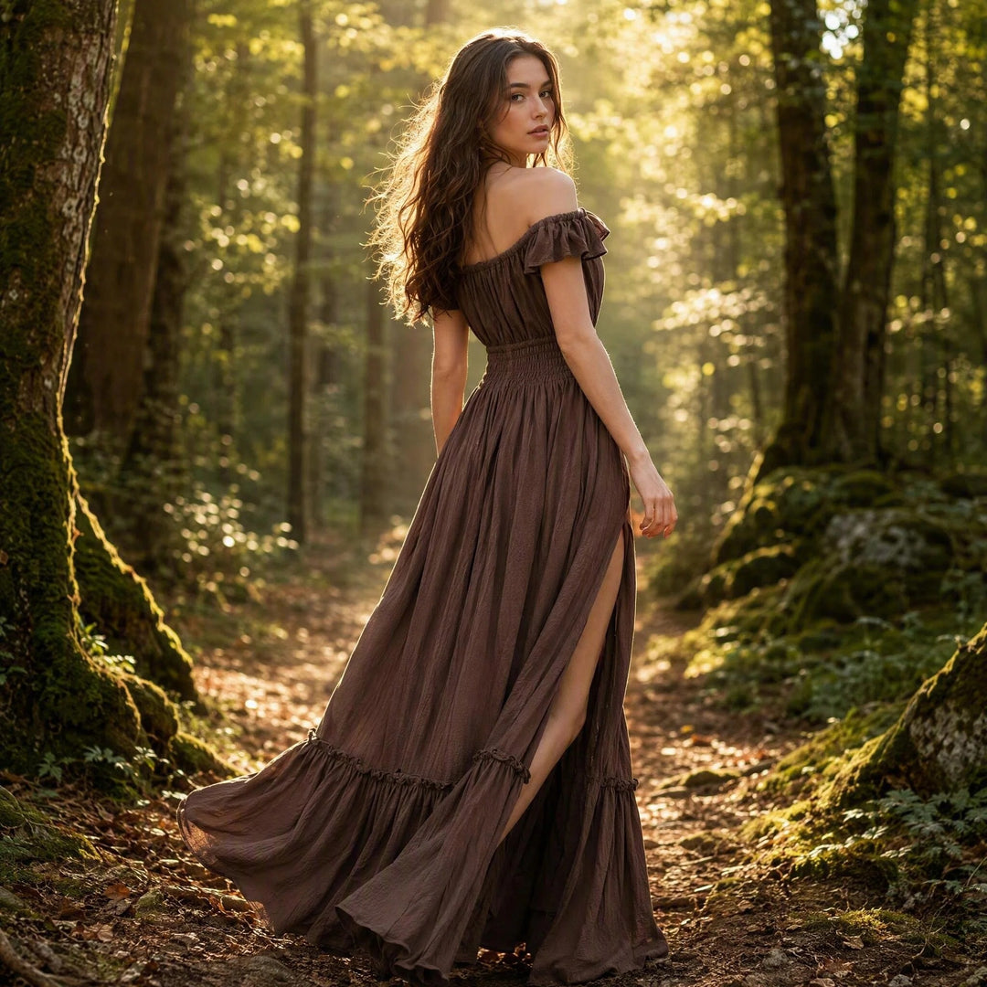 A woman in a coffee off-shoulder gown stands on a forest path; the dress features ruffled sleeves, an elasticated waist, a side slit, and a tiered hem. She turns back sideways.