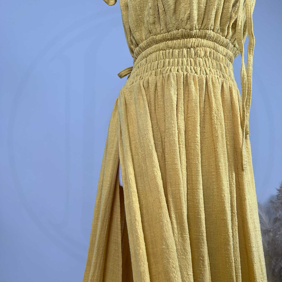 A close-up of a Tethys gown on a mannequin shows a wide elasticated shirred waist, hanging ties, and a side slit in textured fabric.
